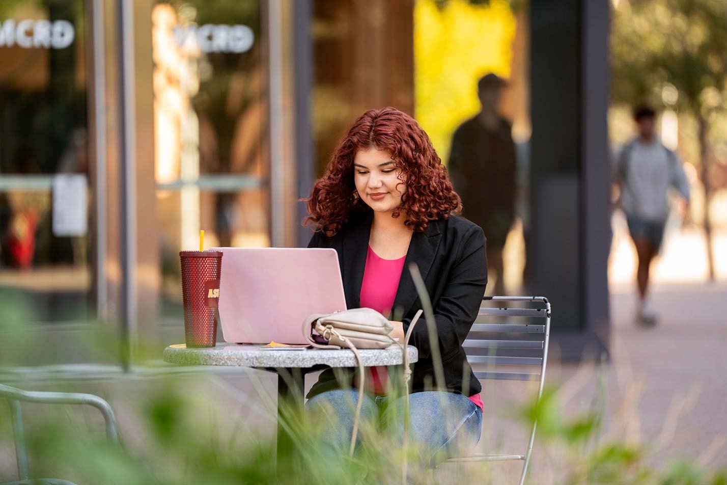 A student in a blazer in ASU's Business (Retail Management) degree studies on a laptop open outside a café ASU's Business (Retail Management) degree.