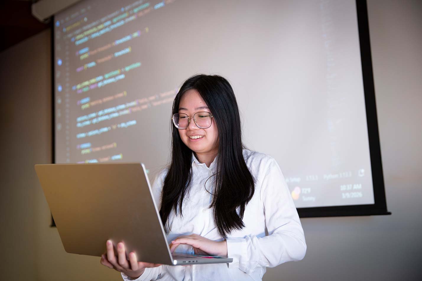 A student smiles while typing, a code-filled screen behind, showcasing expertise gained in ASU's MS in Artificial Intelligence Engineering (Computing Sciences) program.