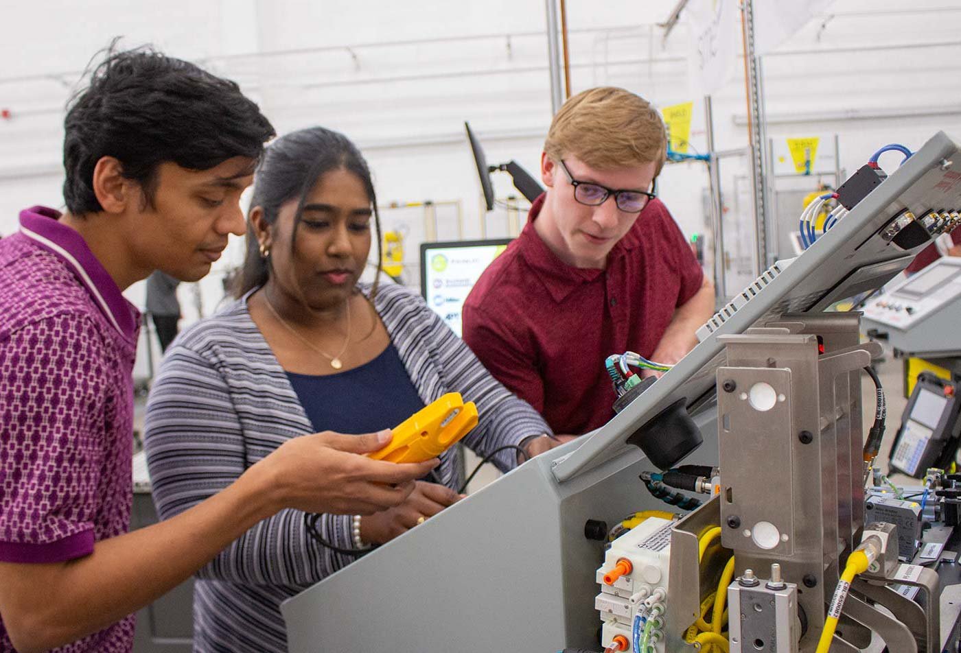 Three students gather around a machine, examining components and using tools, illustrating hands-on learning in ASU's Manufacturing Systems degree.