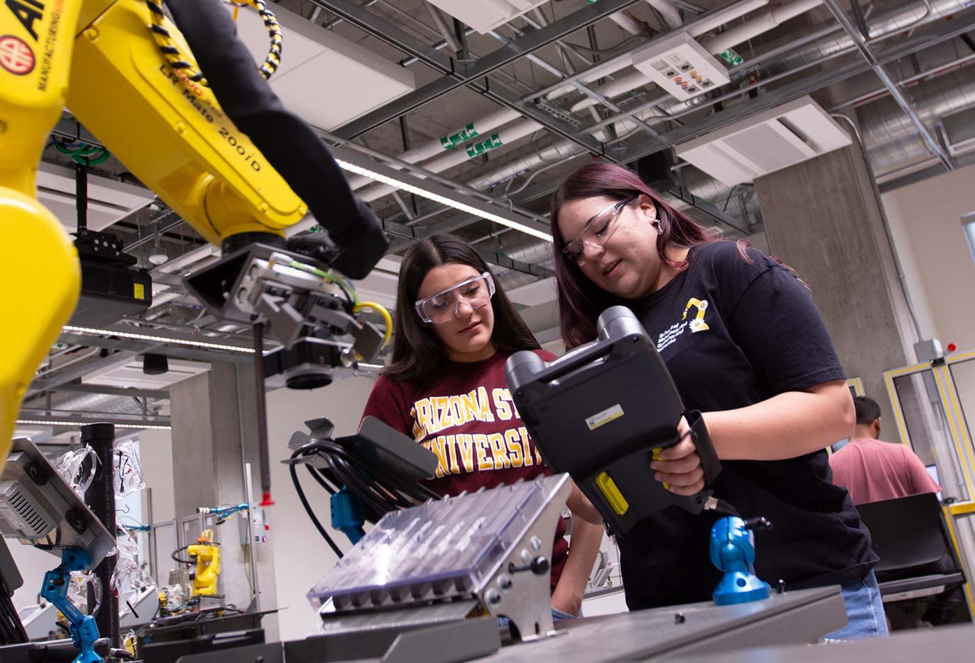 Two students examine a device connected to a robotic arm at a workstation, illustrating applications in ASU's Manufacturing Engineering program.