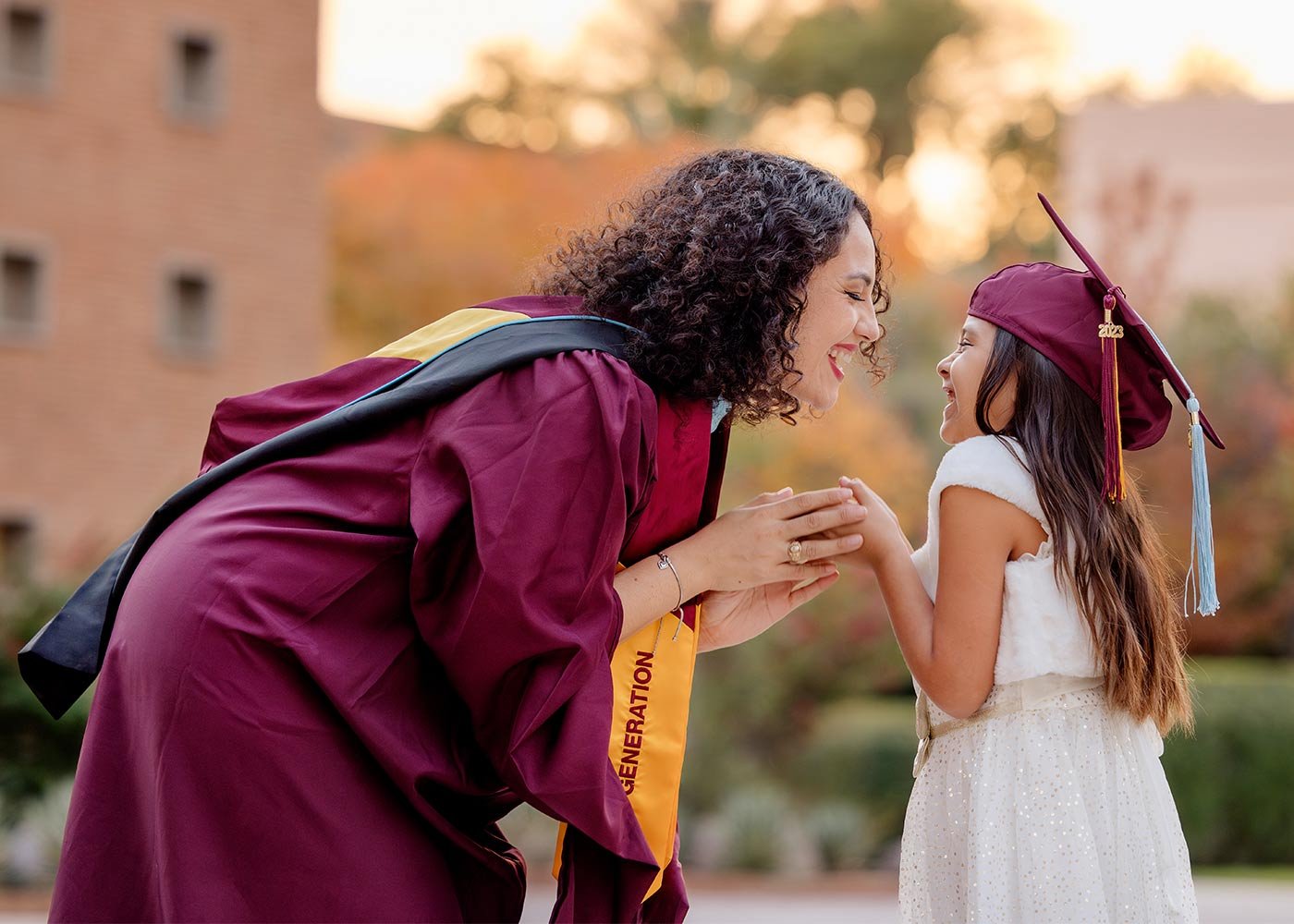 A first-generation master's degree graduate in maroon cap and gown embraces a young girl in a light dress, celebrating achievements in ASU's Advanced Studies program.