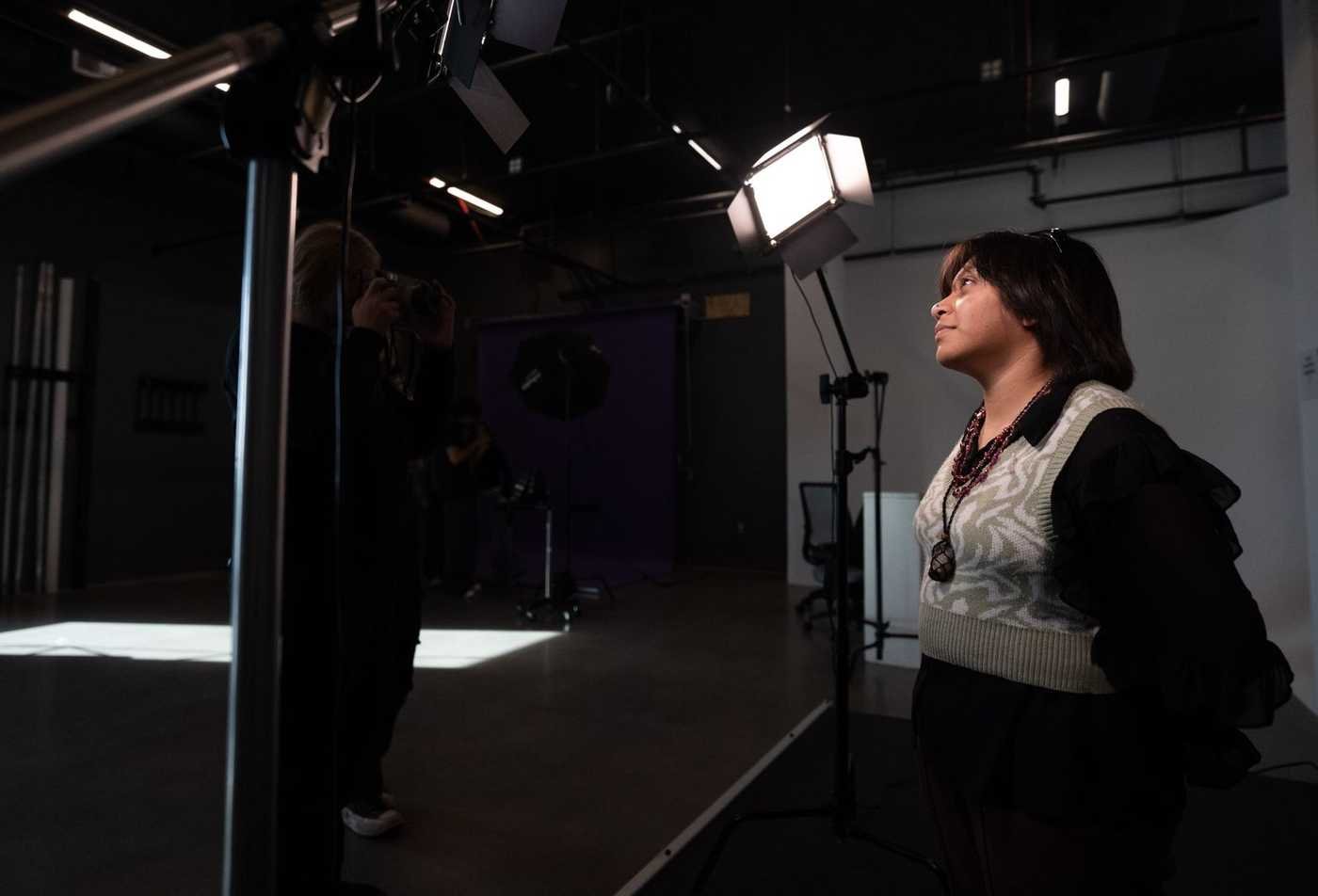 A student poses under lights in the photography lighting studio while another takes a picture, showing the medium explored in the BFA in Art (Photography).