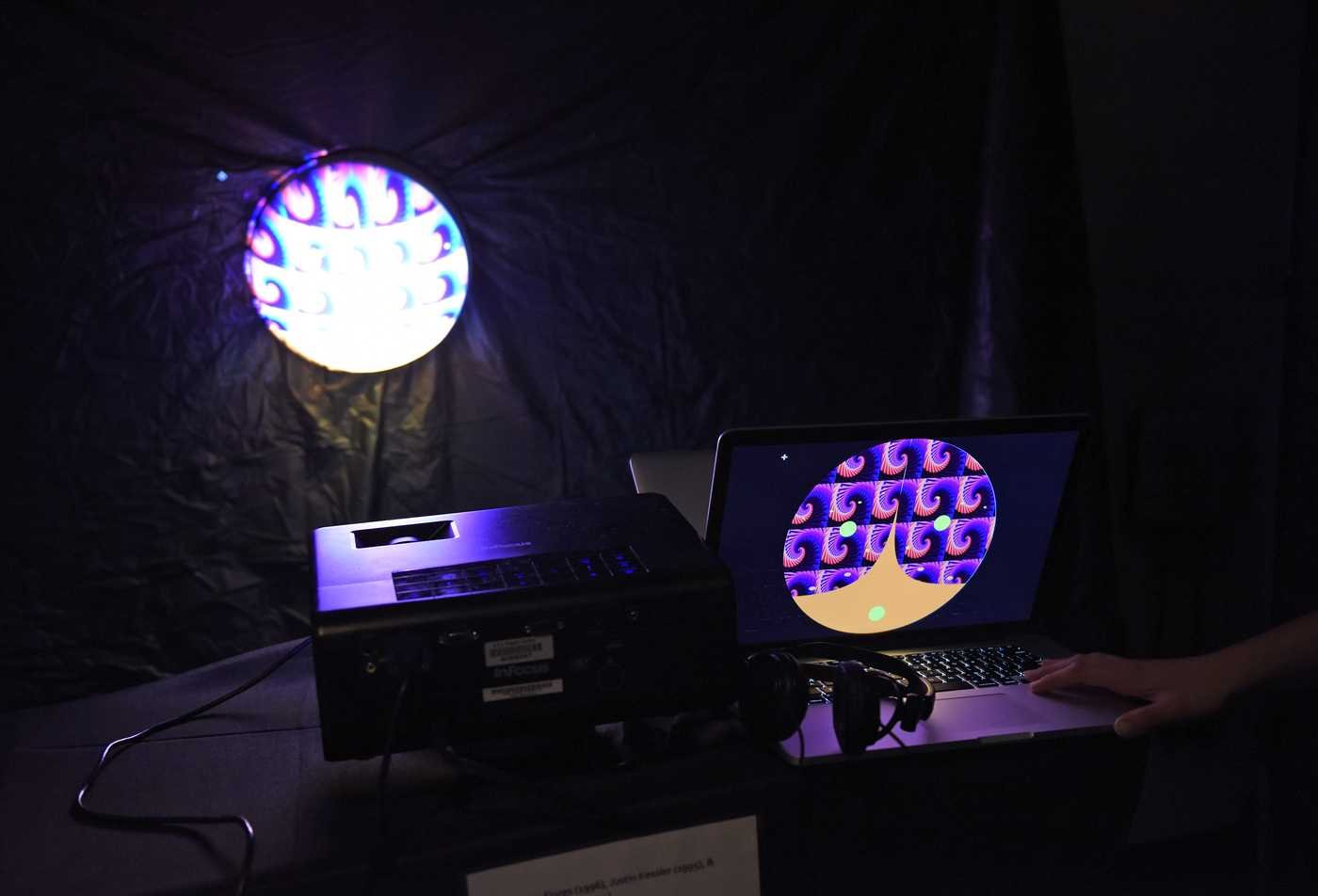 A laptop displaying a colorful circular pattern with a vibrant circular projection on the backdrop demonstrates the field explored in the BA in Media Arts and Sciences.