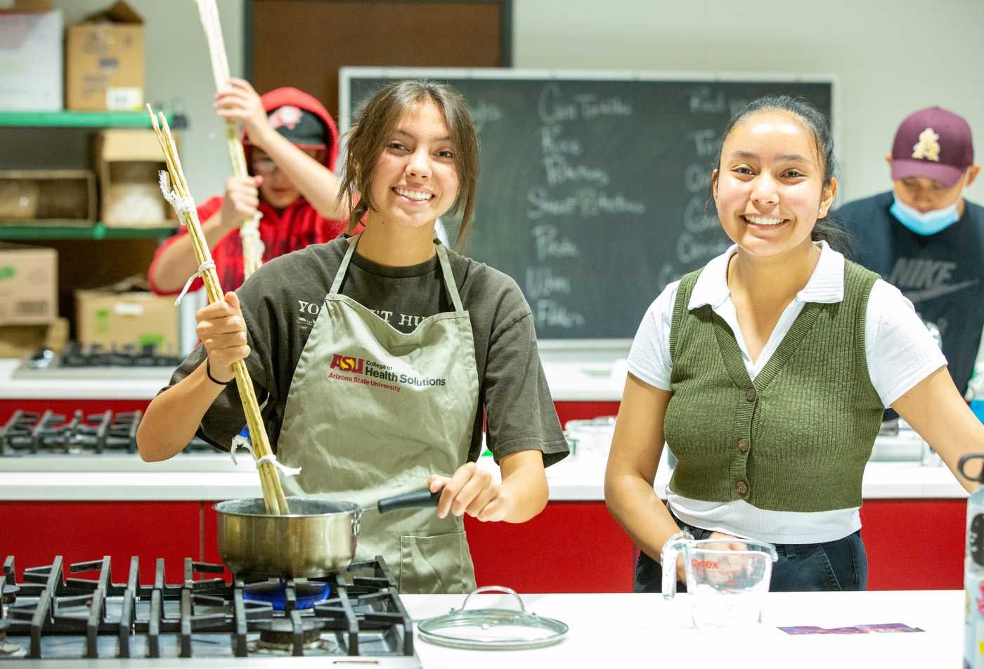 Two students cook in a kitchen, one stirring a pot while another smiles, displaying the skills examined in the BAS in Applied Science (Applied Nutrition and Health)