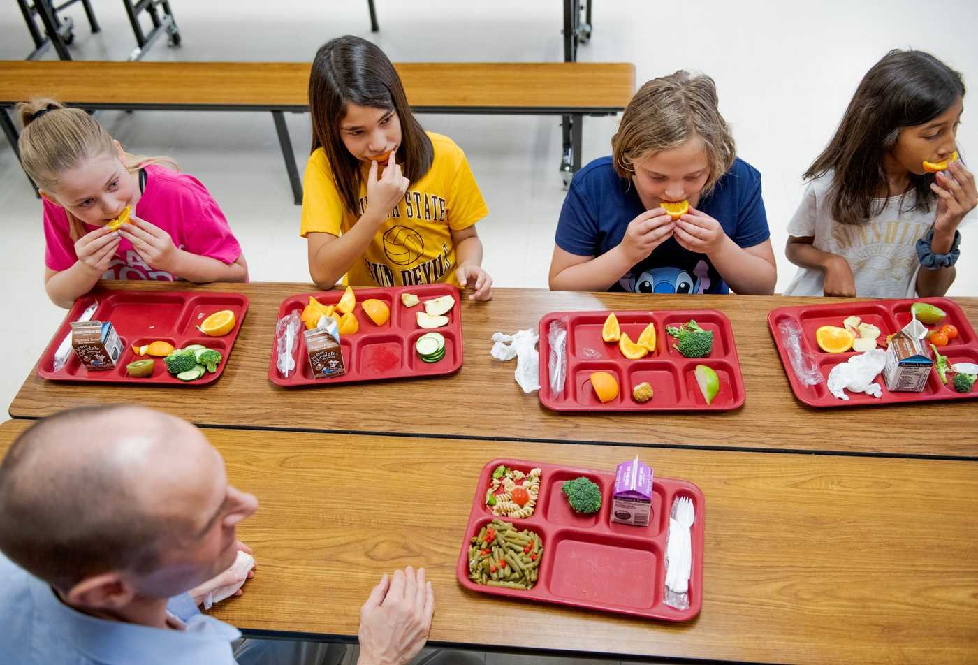 A group of four students at a school cafeteria table eat fruit and vegetables from red trays, demonstrating the field examined in the certificate in School Nutrition.