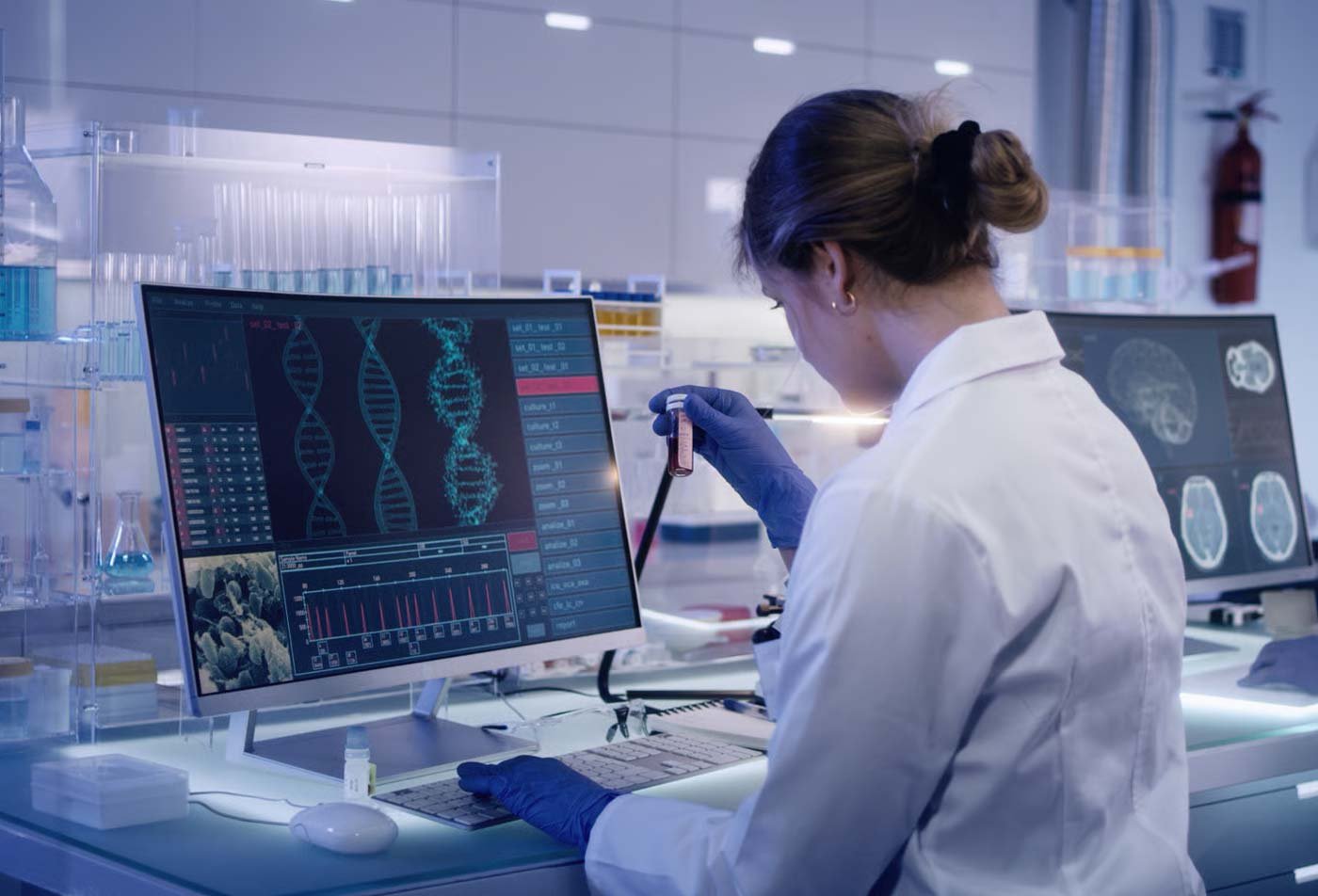 A researcher in a white lab coat holds a test tube in a modern lab, with two computer monitors displaying DNA data and charts, symbolizing the hands-on researched explored in the MS in Biomedical Diagnostics