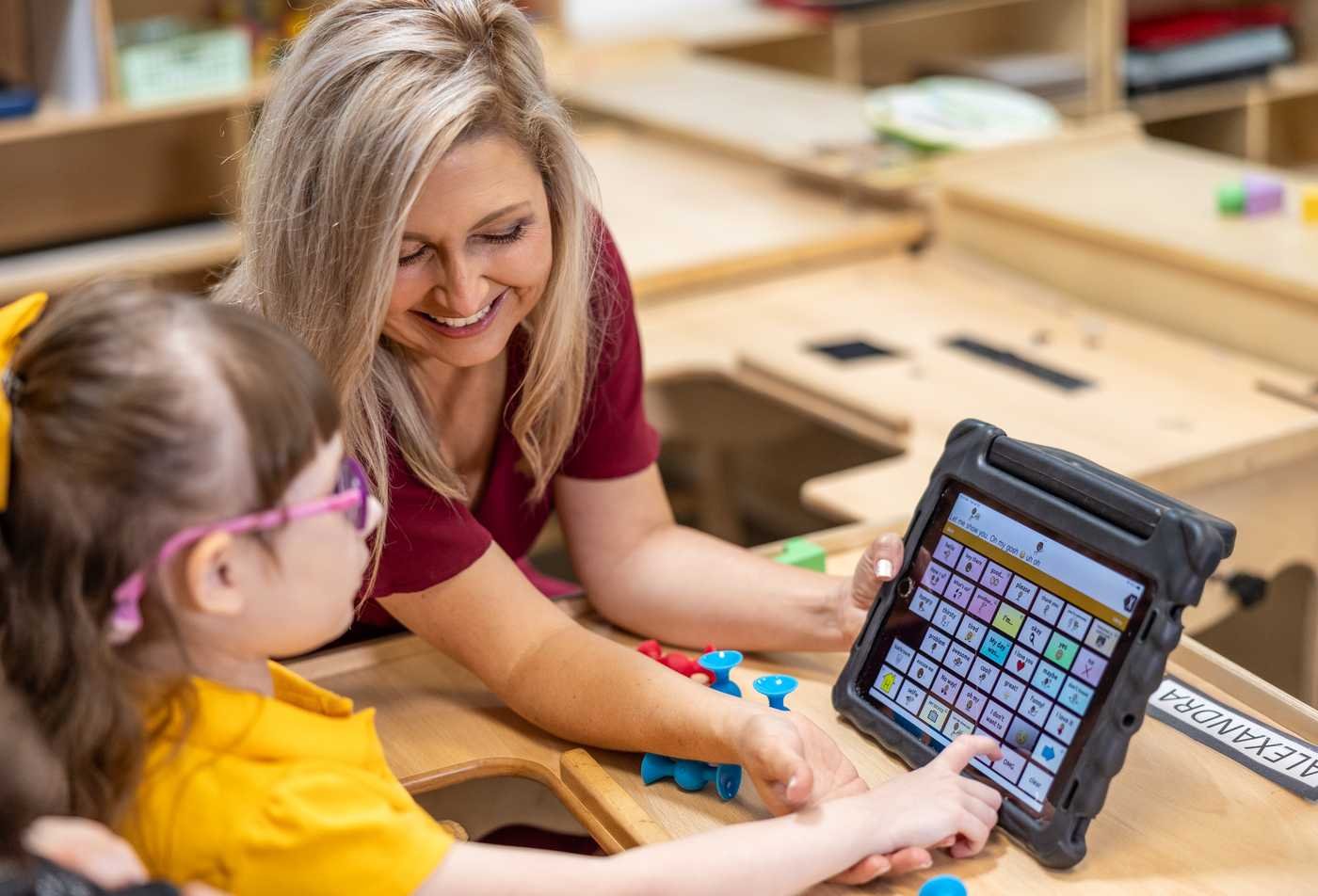 A smiling professional uses a communication device with a young girl, demonstrating the expertise gained in the certificate in Communication Sciences and Disorders
