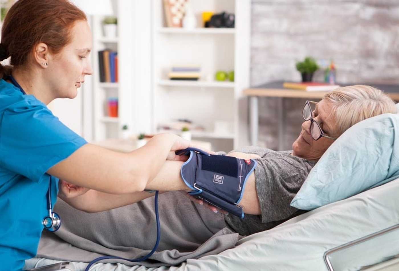 A healthcare professional takes the blood pressure of a patient lying in bed, showcasing a caring interaction in a clinical setting examined in the certificate in Hospice and Palliative Care.