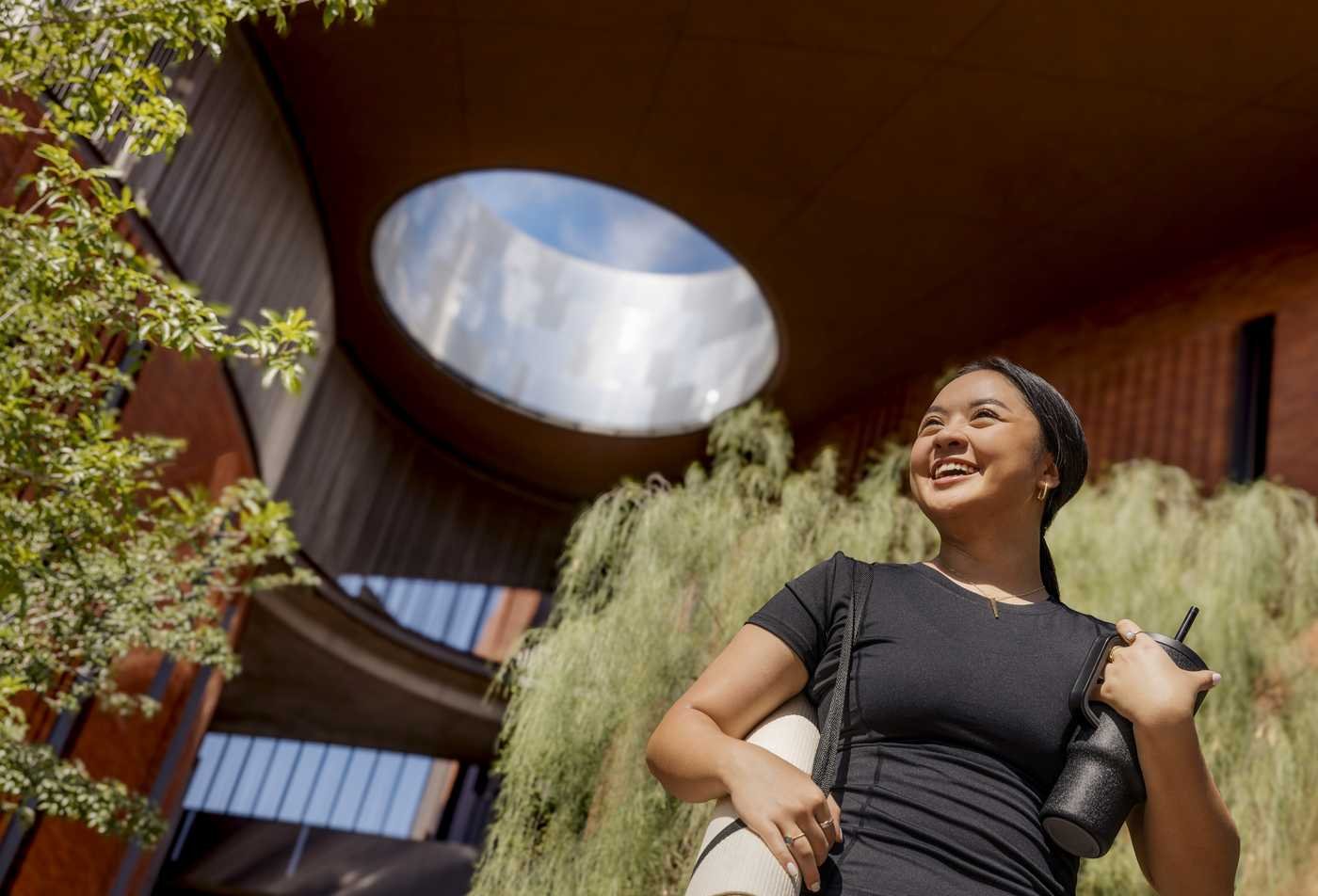 A student with a water bottle and yoga mat stands outside on campus, putting the principles learned in the certificate in Health and Wellness Coach to action.