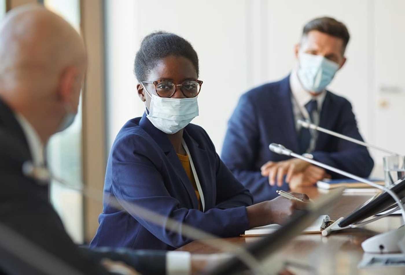 Three individuals in a conference room, wearing face masks, engage in a discussion at a long wooden table, showcasing the principles explored in the BIPH in International Health Management.