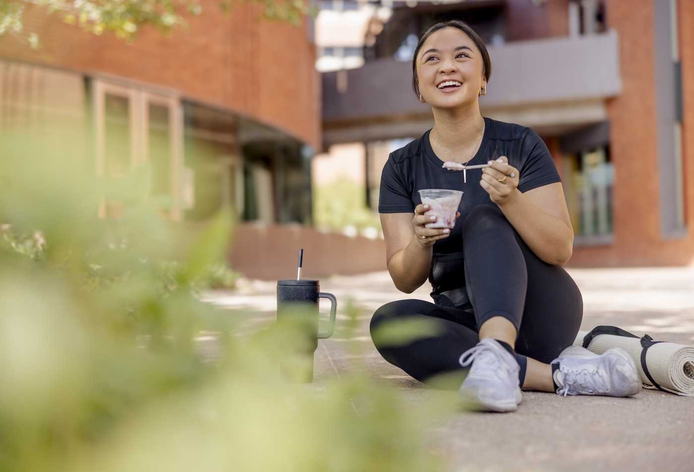 A students sits outdoors, smiling while holding a healthy yogurt snack after a workout, demonstrating the principles explored in the MS in Medical Nutrition.