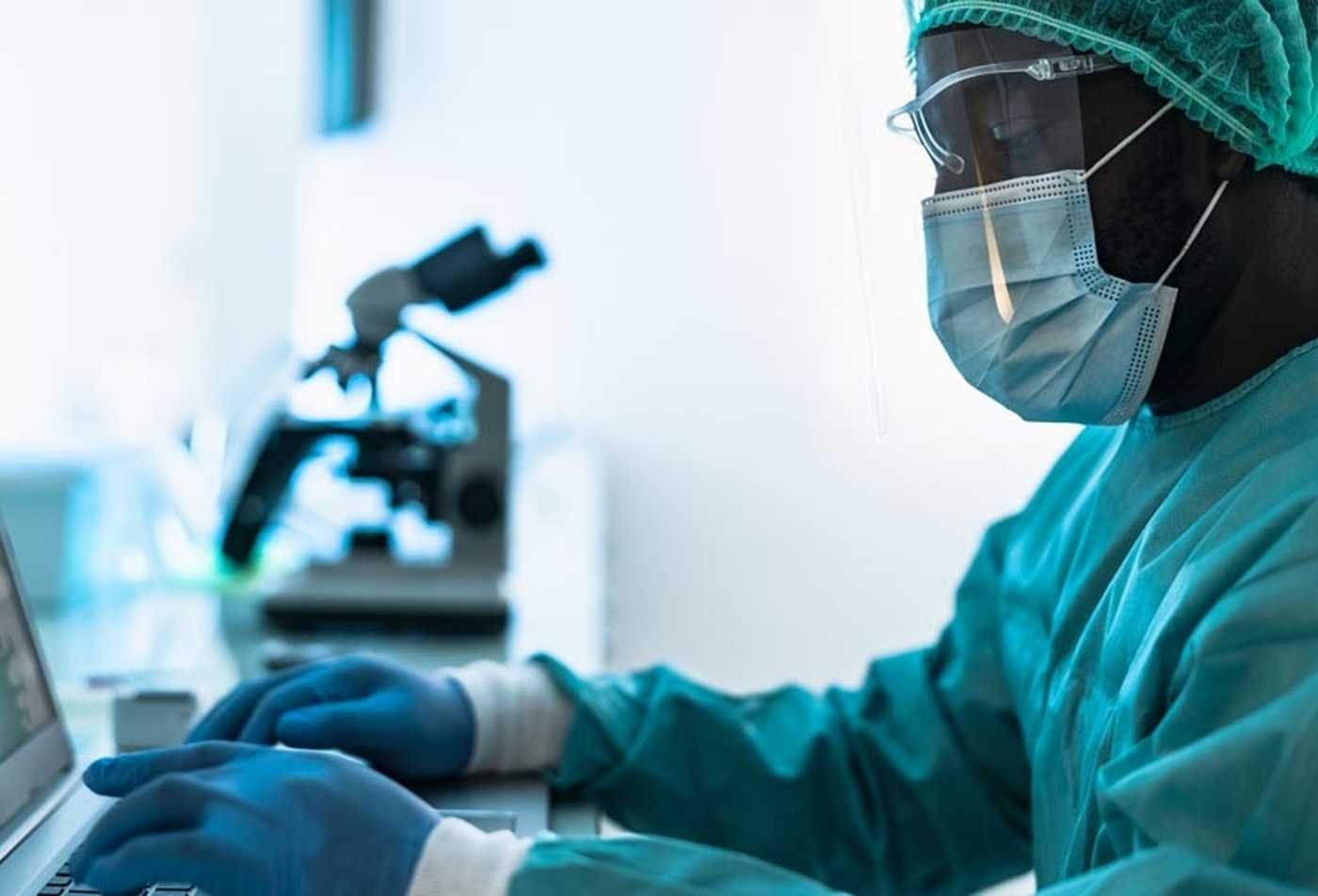 A researcher in gloves and a mask works on a laptop in a lab, highlighting the field explored in the BAS in Applied Science (Medical Laboratory Science)