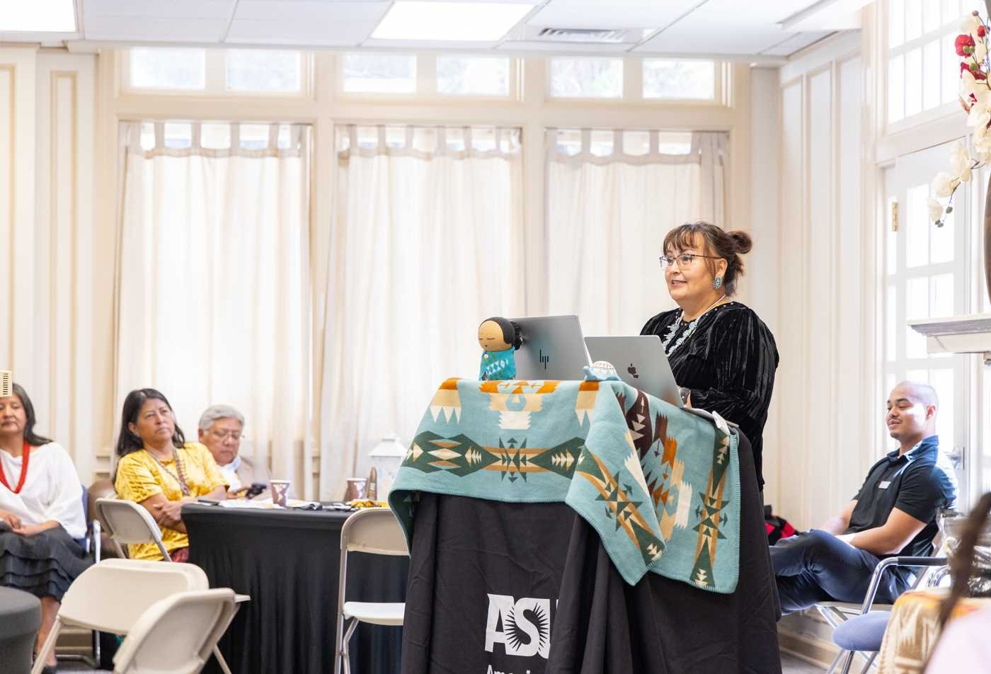 A woman speaks at a podium draped with a Native American-patterned blanket, showcasing the field explored in the certificate in American Indian Nation Governance.