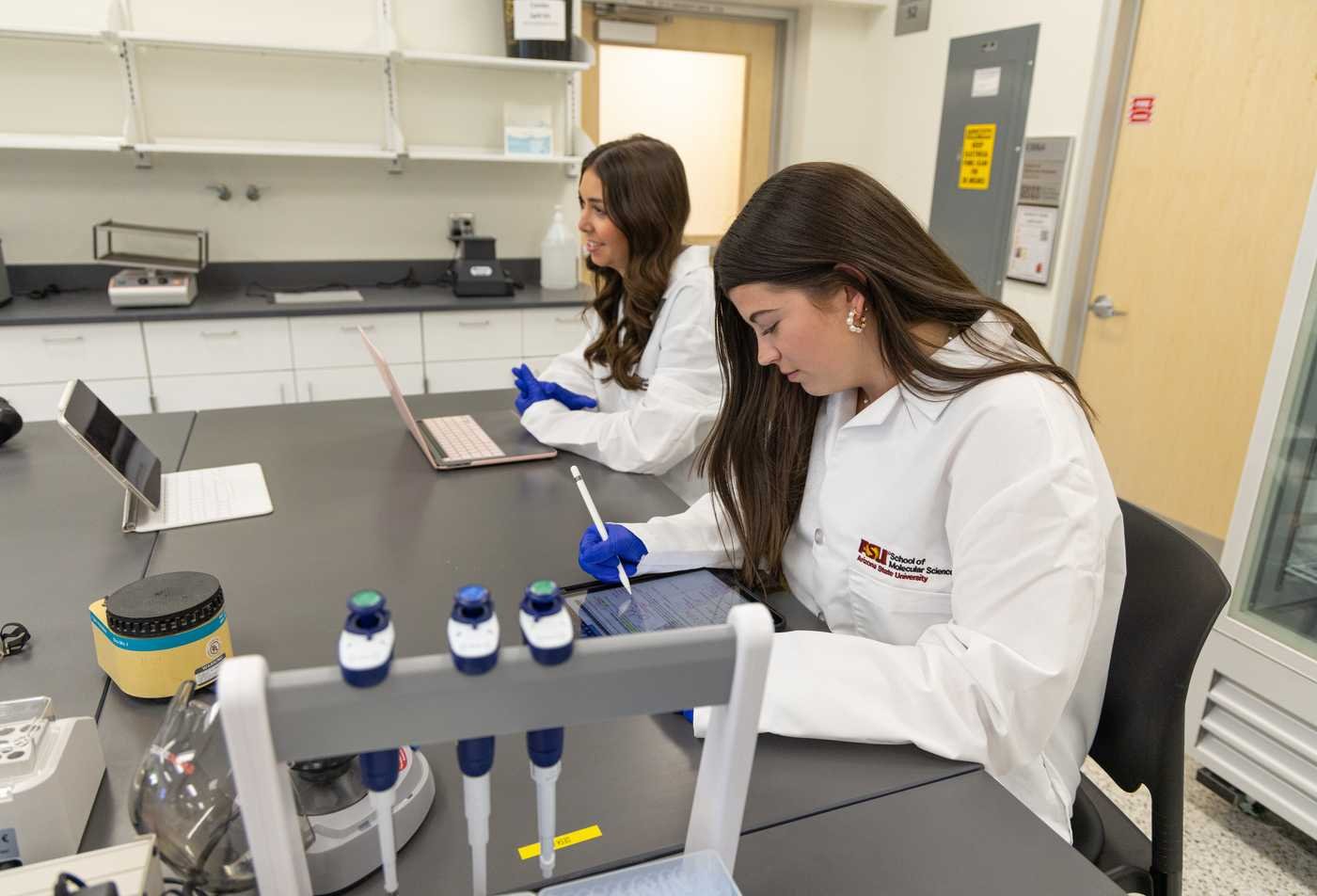 Two students take notes in a lab with ASU white coats on, demonstrating hands-on learning in the MS in Biochemistry.