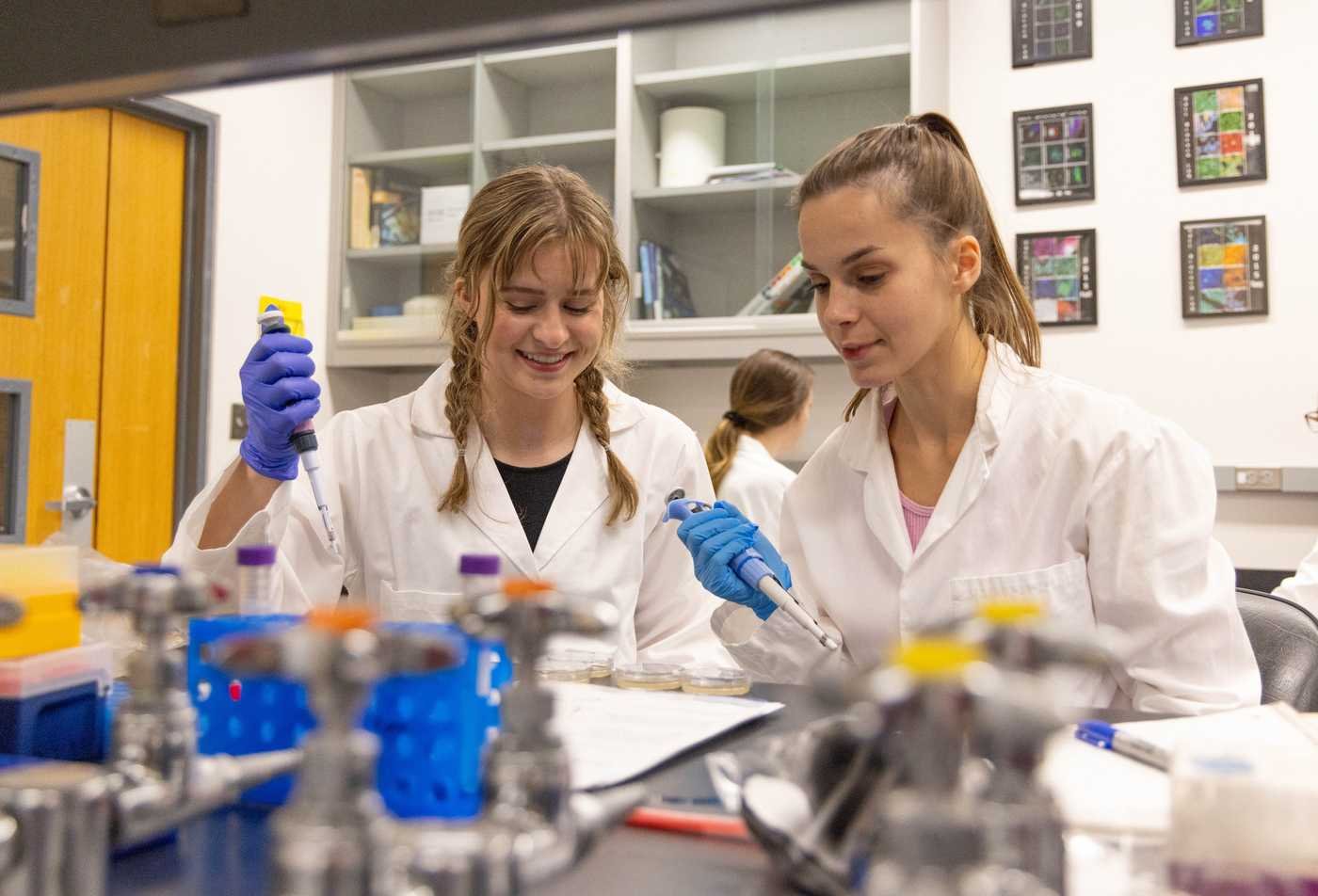Two students wearing science glasses and lab coats work on an experiment, reflecting hands-on research in the Molecular and Cellular Biology, MS.