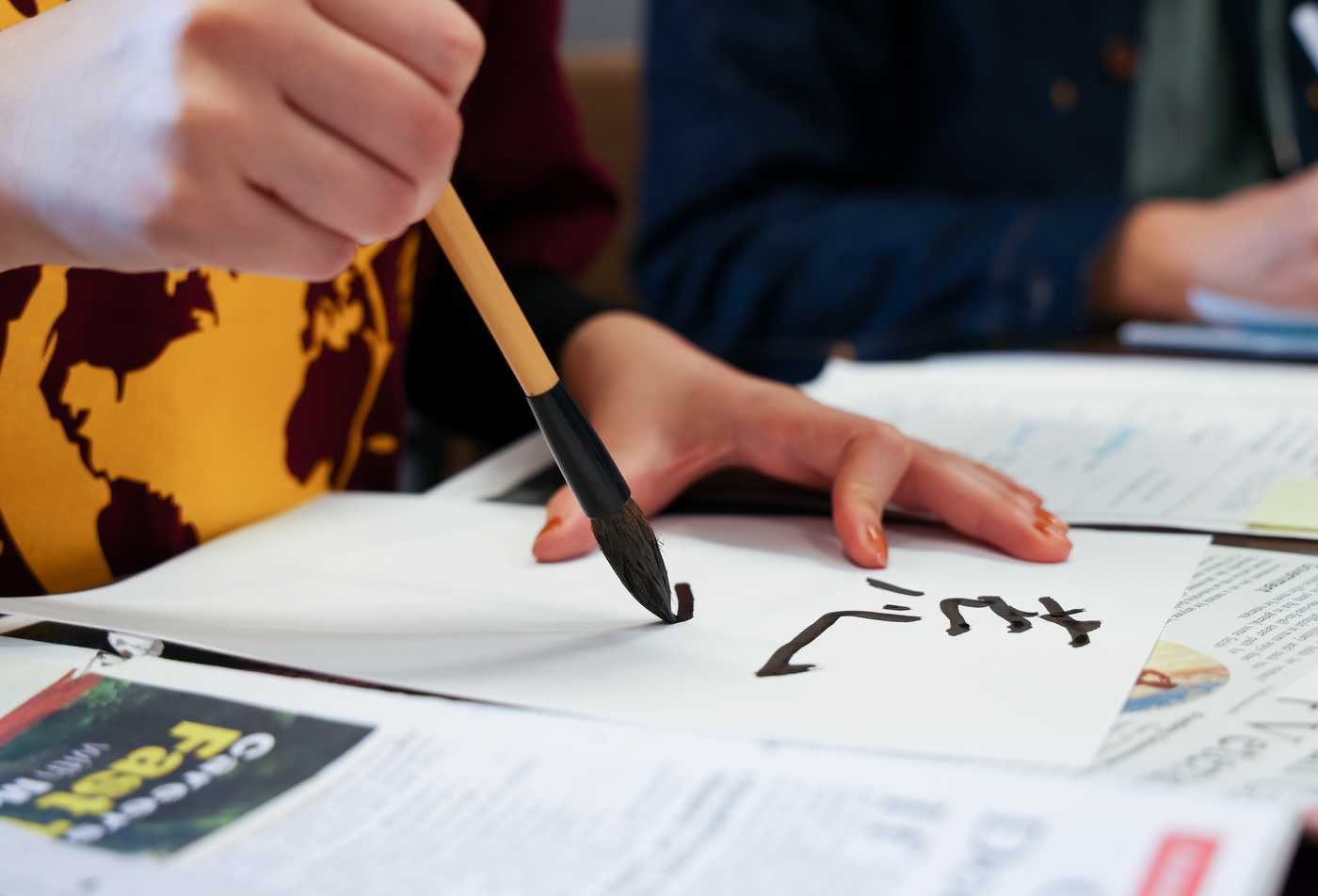 A female student practices brush calligraphy on a sheet of paper while seated at a table with textbooks and class materials, demonstrating hands-on learning in the minor in Asian Languages - Chinese.