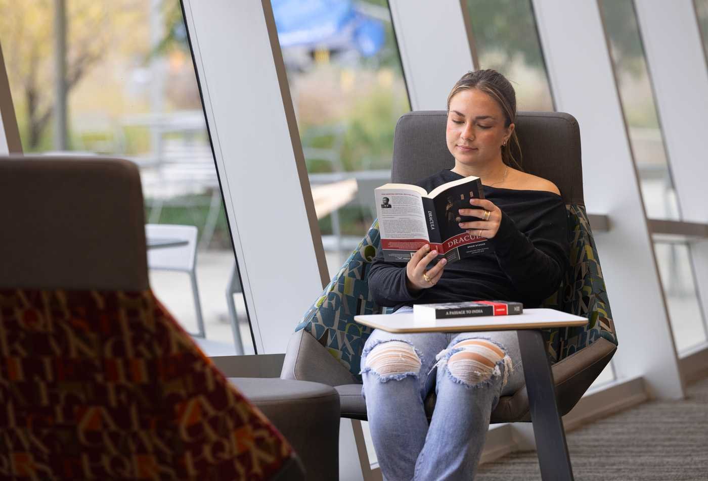 A student sits in a cushioned chair in a sunlit campus lounge, reading a copy of Dracula by Bram Stoker, showcasing the field explored in the minor in Classics.