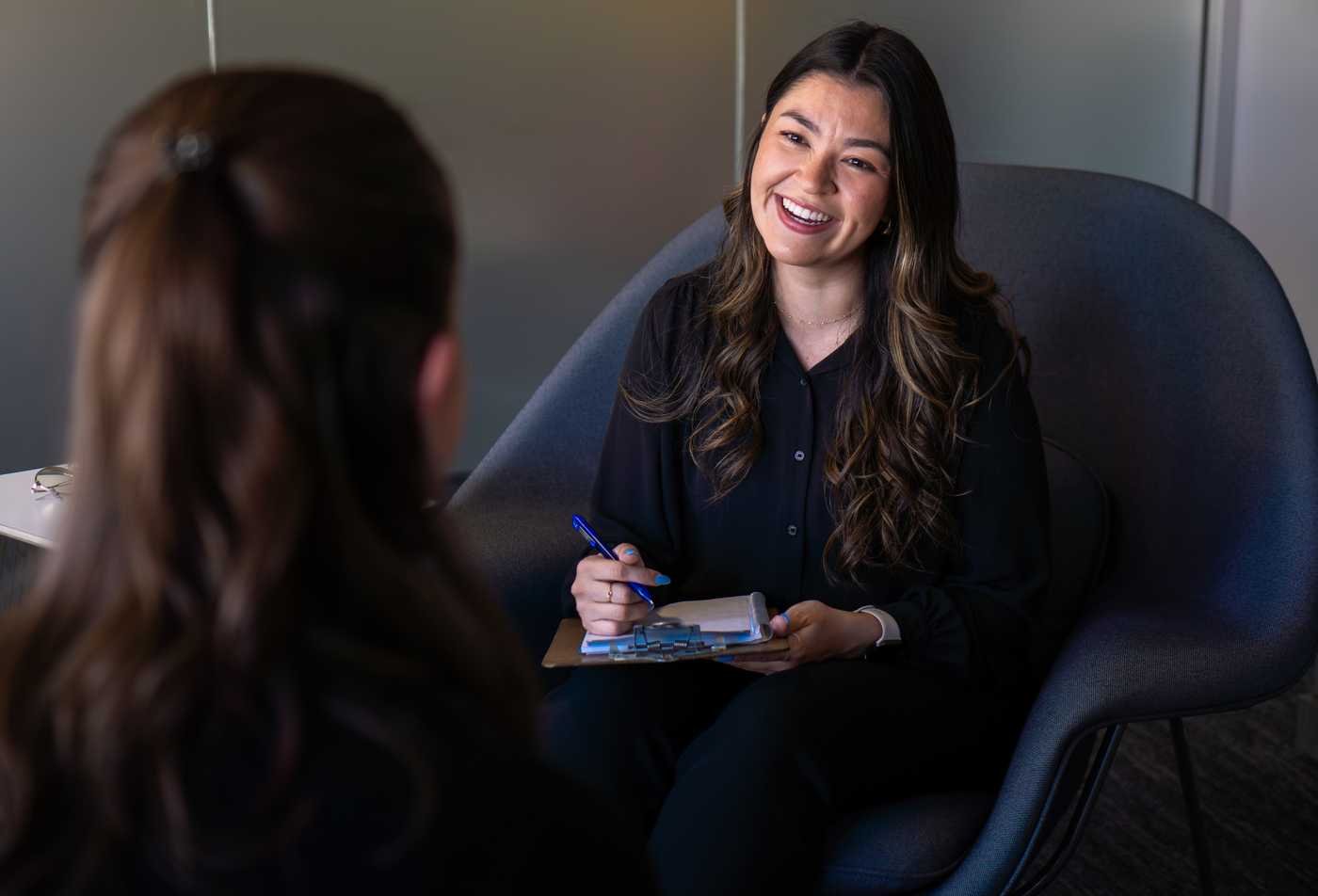 Psychology student Karen Aguilar smiles while holding a clipboard and talking with another person in an office, showcasing the expertise gained in the MS in Clinical Psychology.