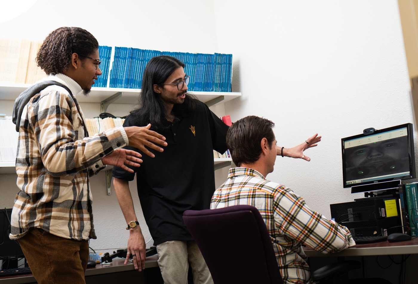 Three ASU students collaborate in a research lab, with two standing and one seated at a desktop computer with software