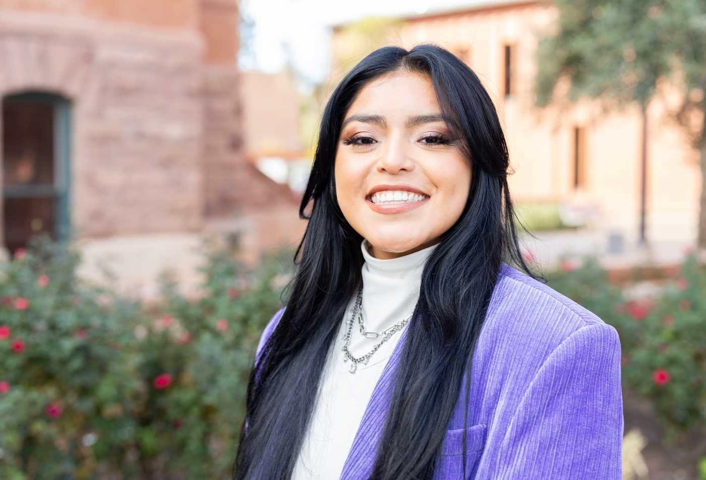 A female ASU student wearing a purple blazer smiles brightly into the camera.