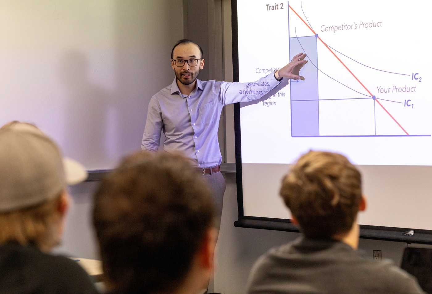 An instructor gestures toward a projected economics slide while facing a classroom of seated students, reflecting practical learning in the minor in Economics.