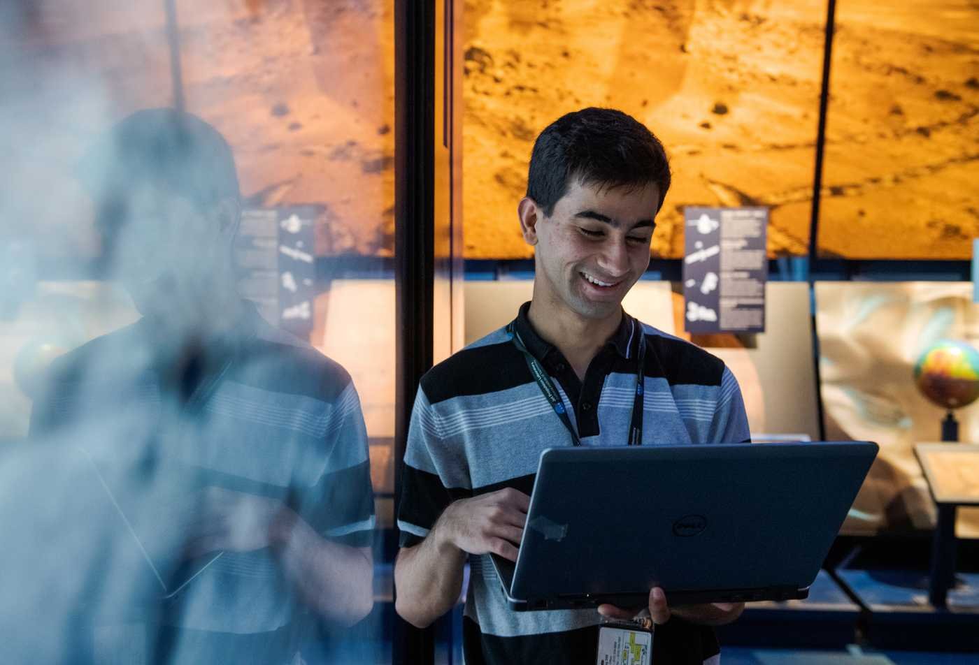 A student smiles while looking at a tablet in front of a projected image of Mars, symbolizing the field exploired in the Exploration Systems Design (Instrumentation), PhD.