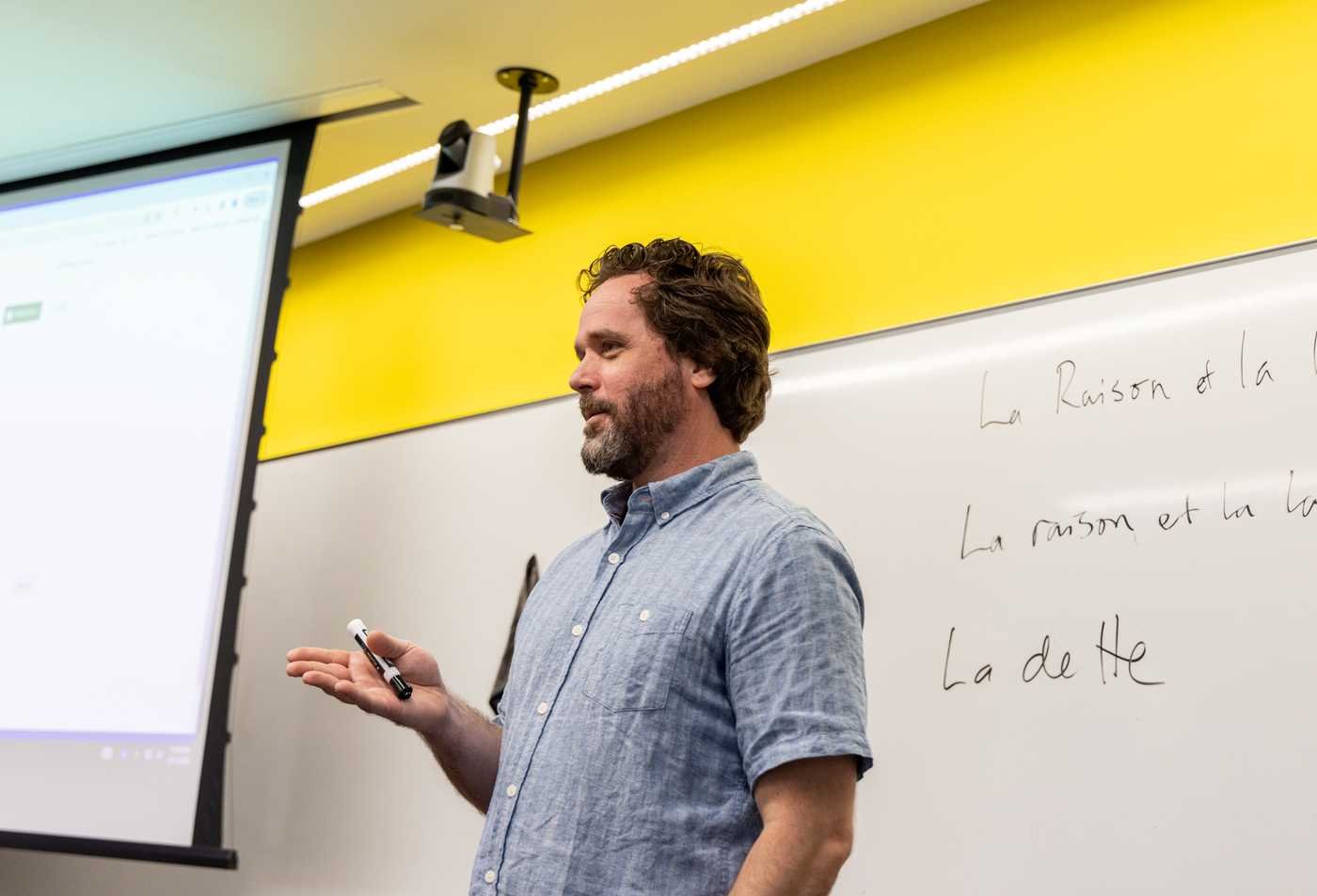 French professor stands in front of a white board with writing, demonstrating the field explored in the French Linguistics, MA.