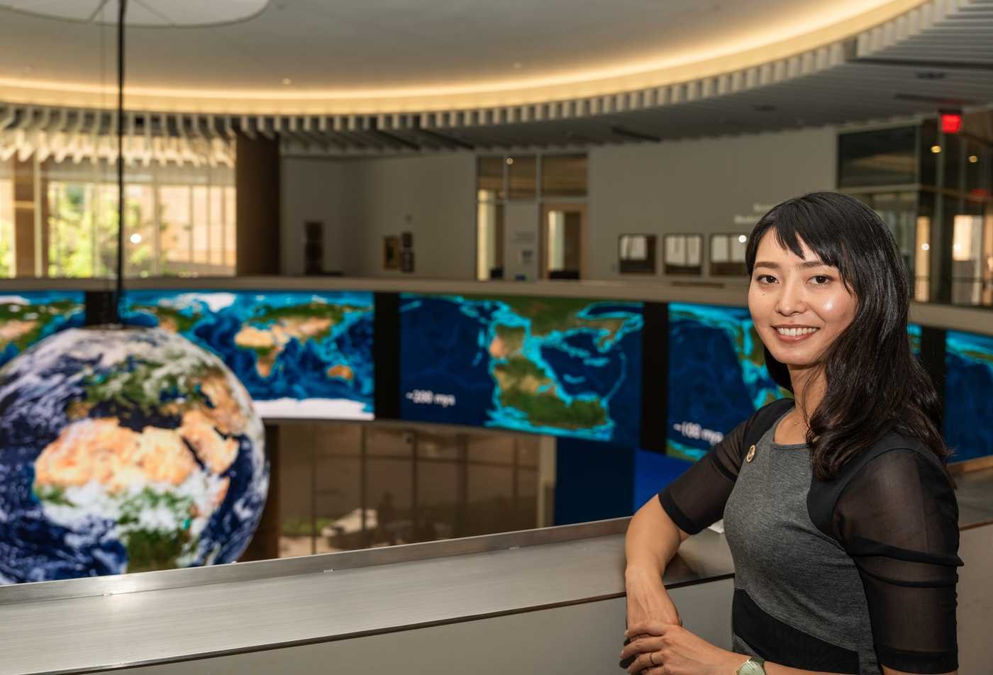 A student poses in front of screens displaying the globe, symbolizing the field explored in the Global Security, MA.