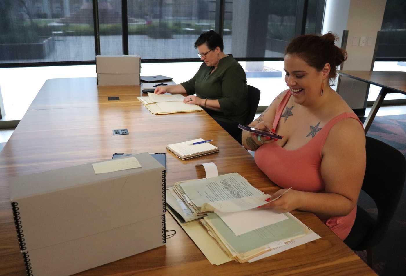 Two researchers sit at a long wooden table in an archive reading room, reviewing historical documents and folders from archival boxes, demonstrating hands-on learning in the minor in History.