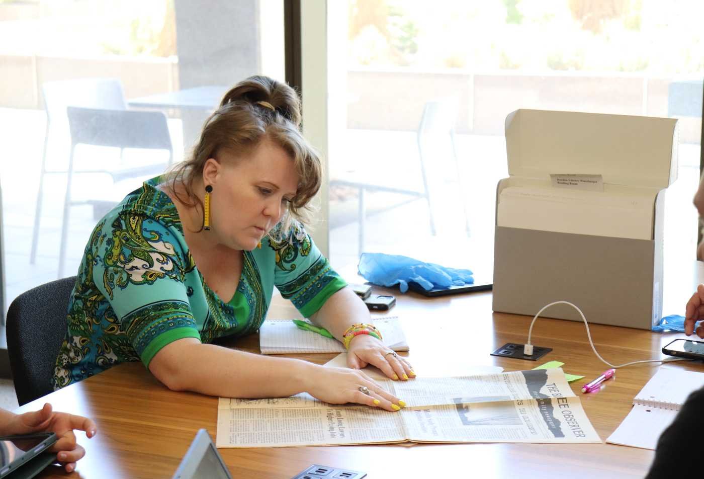 A student looks at a historical newspaper called "The Observer" at a library reading room table, surrounded by archival materials, showcasing hands-on learning in the History, MA.