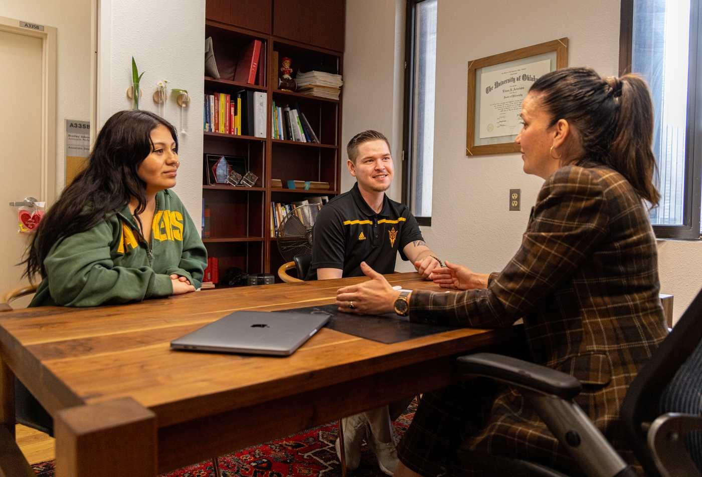 Two ASU students meet with a faculty member in her office, with bookshelves and diploma visible on the wall behind them, showcasing the collaborative setting of the PhD in Comparative Culture and Language.