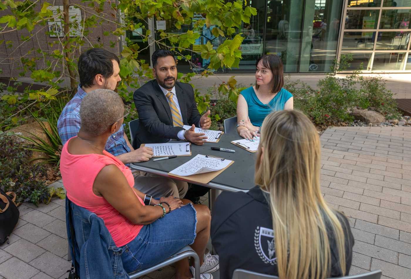 Two male and three female graduates sit at a table outdoors. They engage in conversation and on the table are clipboards with writing.
