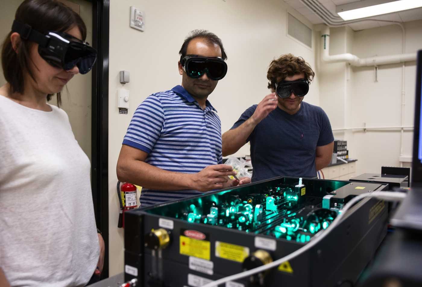 Students and professor look at a physics lazer research machine, demonstrating hands-on learning in the Physics, PhD