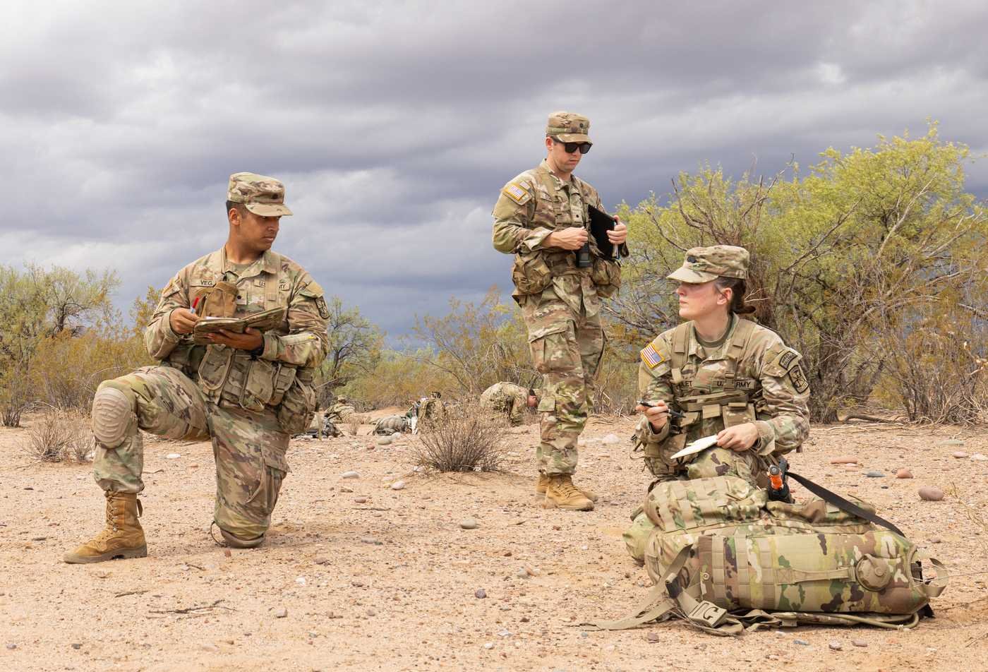 Three students in camouflage uniforms review notes and equipment during an outdoor field training exercise in a desert landscape, demonstrating hands-on learning in the certificate in Working with Military Families.