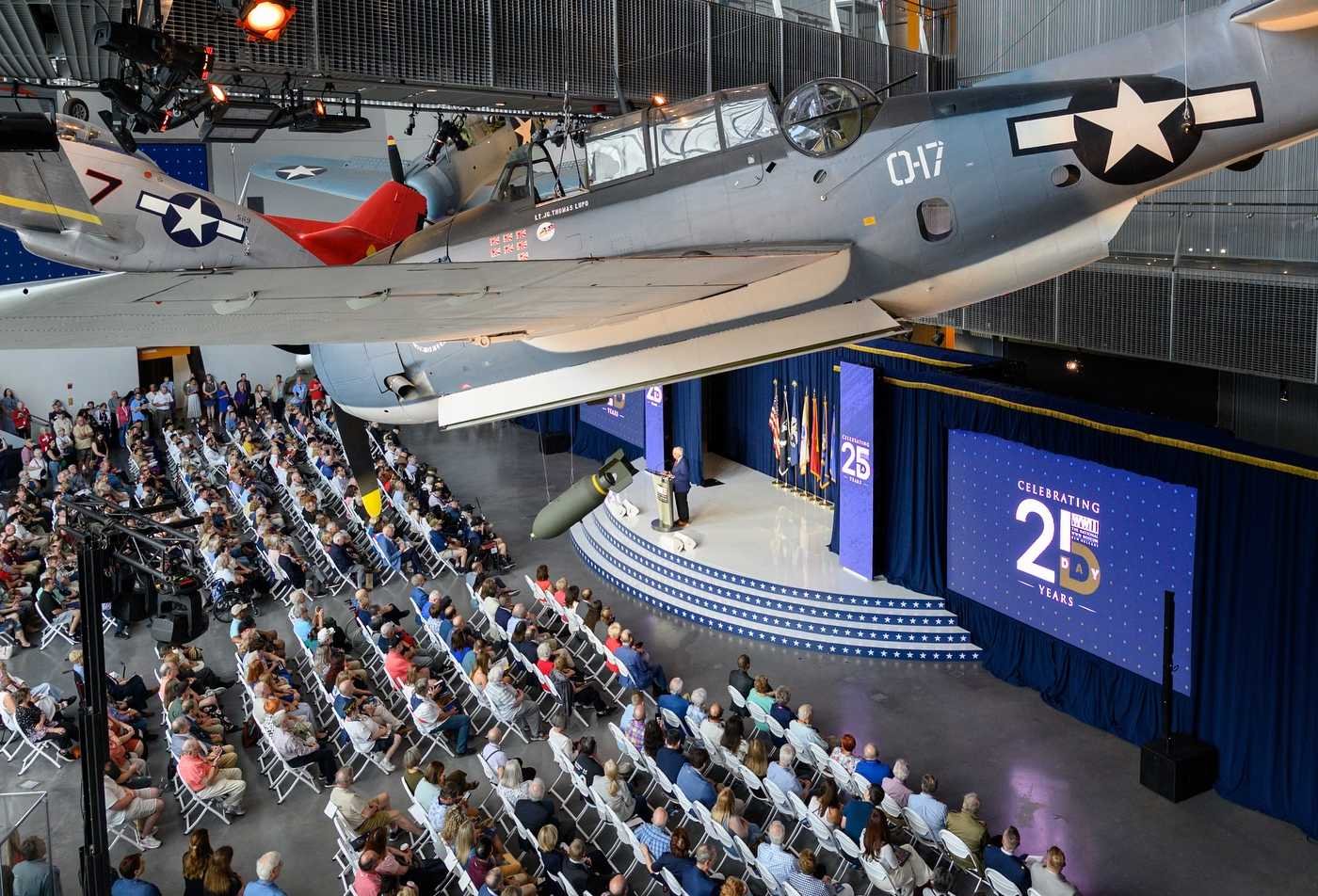 A WWII aircraft hang suspended from the ceiling above attendees seated in rows of white chairs, showcasing the field explored in the World War II Studies, MA.