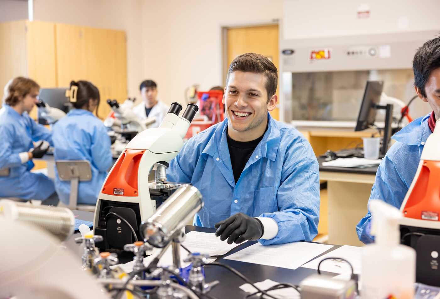 Students in a lab wearing blue lab coats work with microscopes in ASU’s Applied Quantitative Science degree