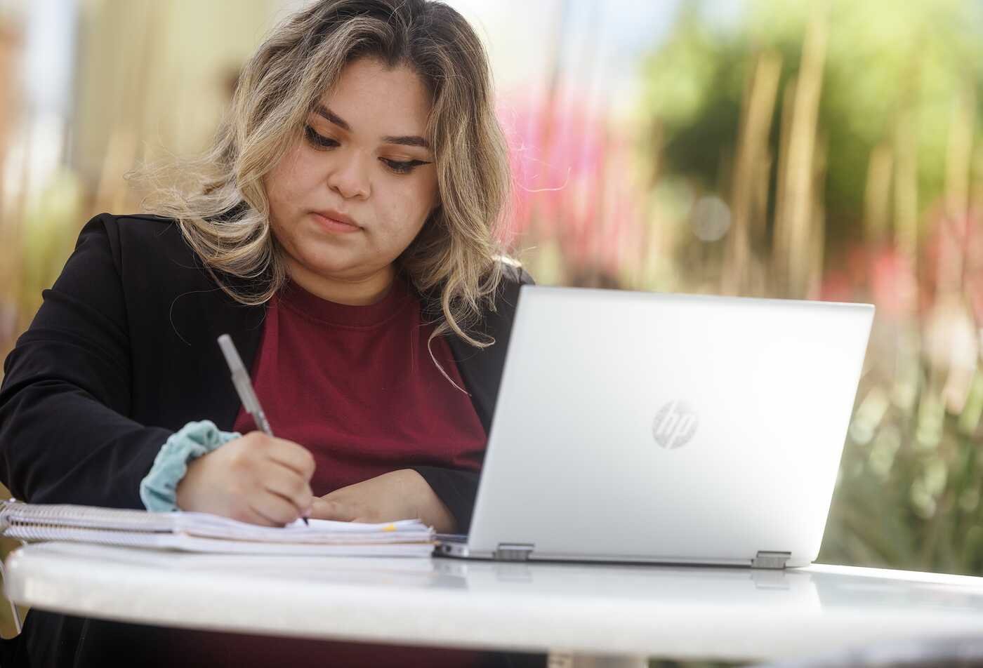 A student writes notes beside a laptop outdoors, developing written communication skills in ASU's Writing certificate.