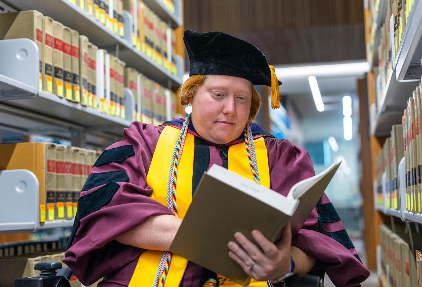 A graduate in academic regalia reads in the Law library, showcasing students' dedication to knowledge and learning in ASU's JD program.