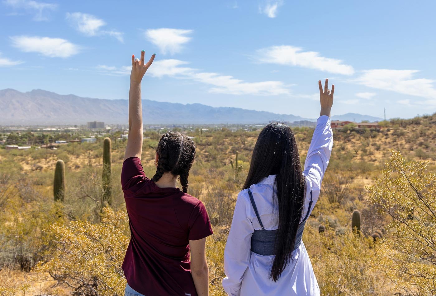 Two students raise their hands in celebration against a desert mountain backdrop, marking their journey through the Tourism concentration.