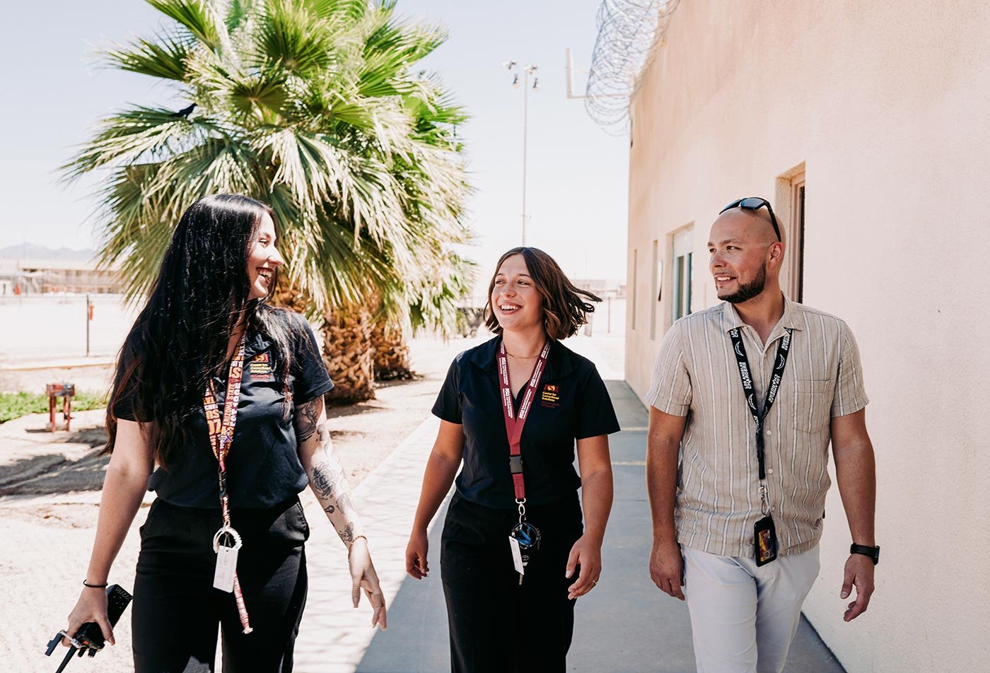 Three students with lanyards chat while walking past a palm tree on campus, reflecting daily life in ASU's Criminology and Criminal Justice program.