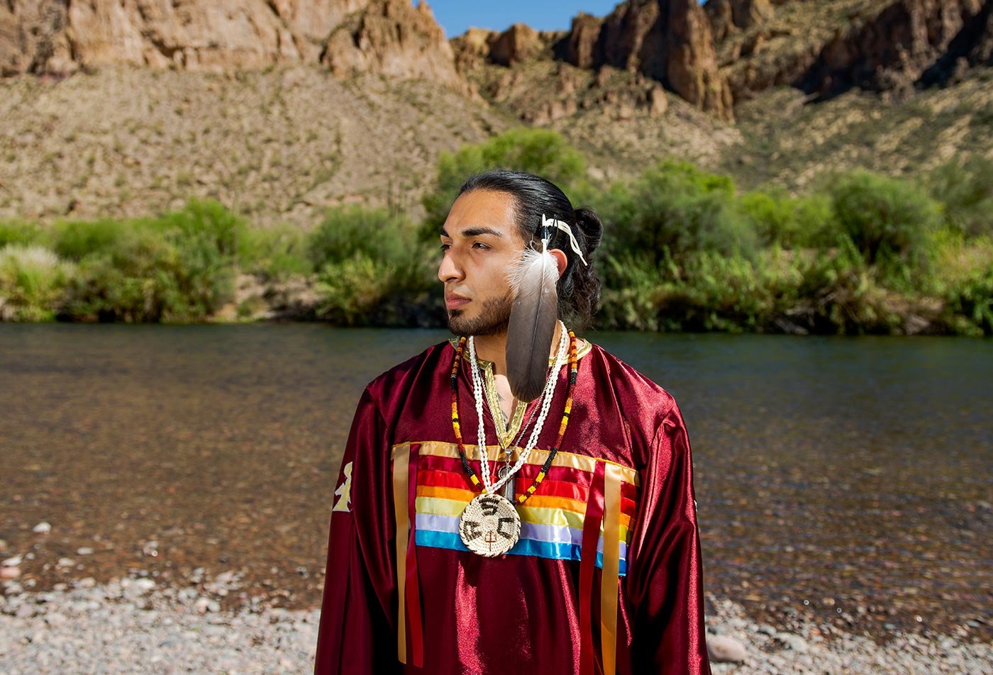 A student in traditional garment and jewelry stands by a river amid rugged mountains, reflecting ASU's American Indian Studies path.