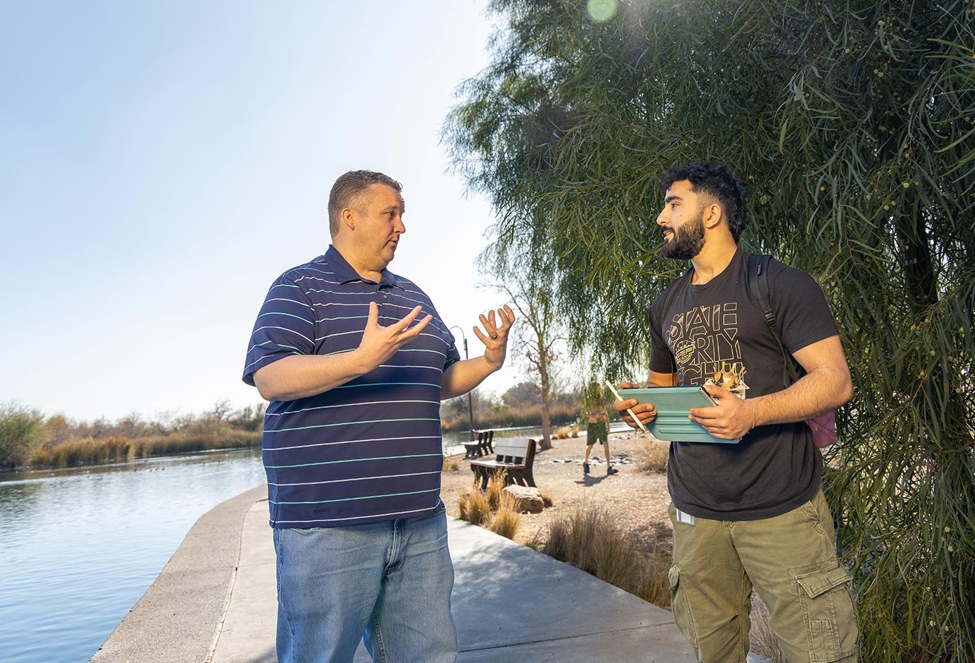 Two men talk beside a calm waterway, reflecting the collaborative learning within ASU's Sustainability concentration.
