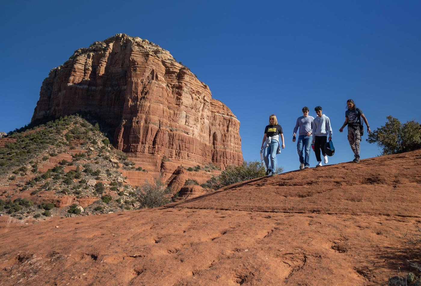 Four students walk across red rock terrain under a clear sky, embodying the spirit of ASU's Sustainable Tourism minor.