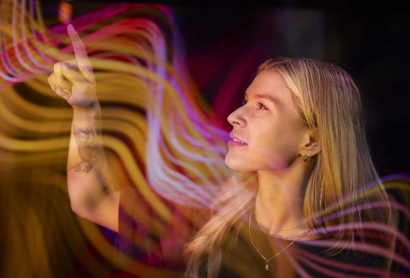 A student points upward, her face illuminated against a colorful swirling background of purple, orange, and yellow that symbolize complex ideas mastered in the MA in Learning Science.
