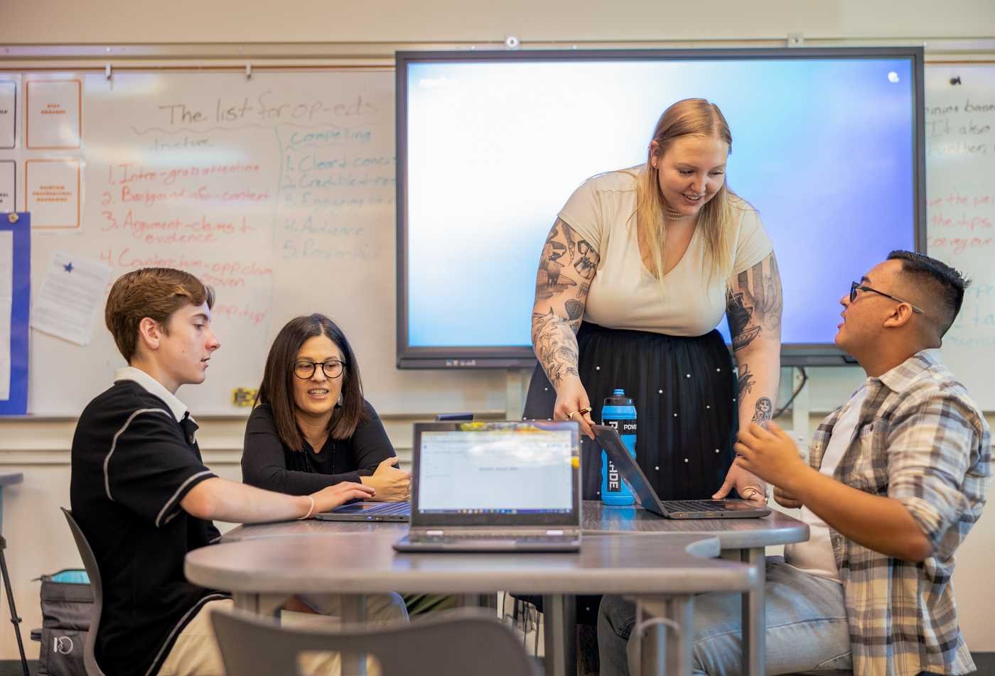 An instructor assists a group of students with a laptop, demonstrating the knowledge gained in the PhD in Educational Technology.