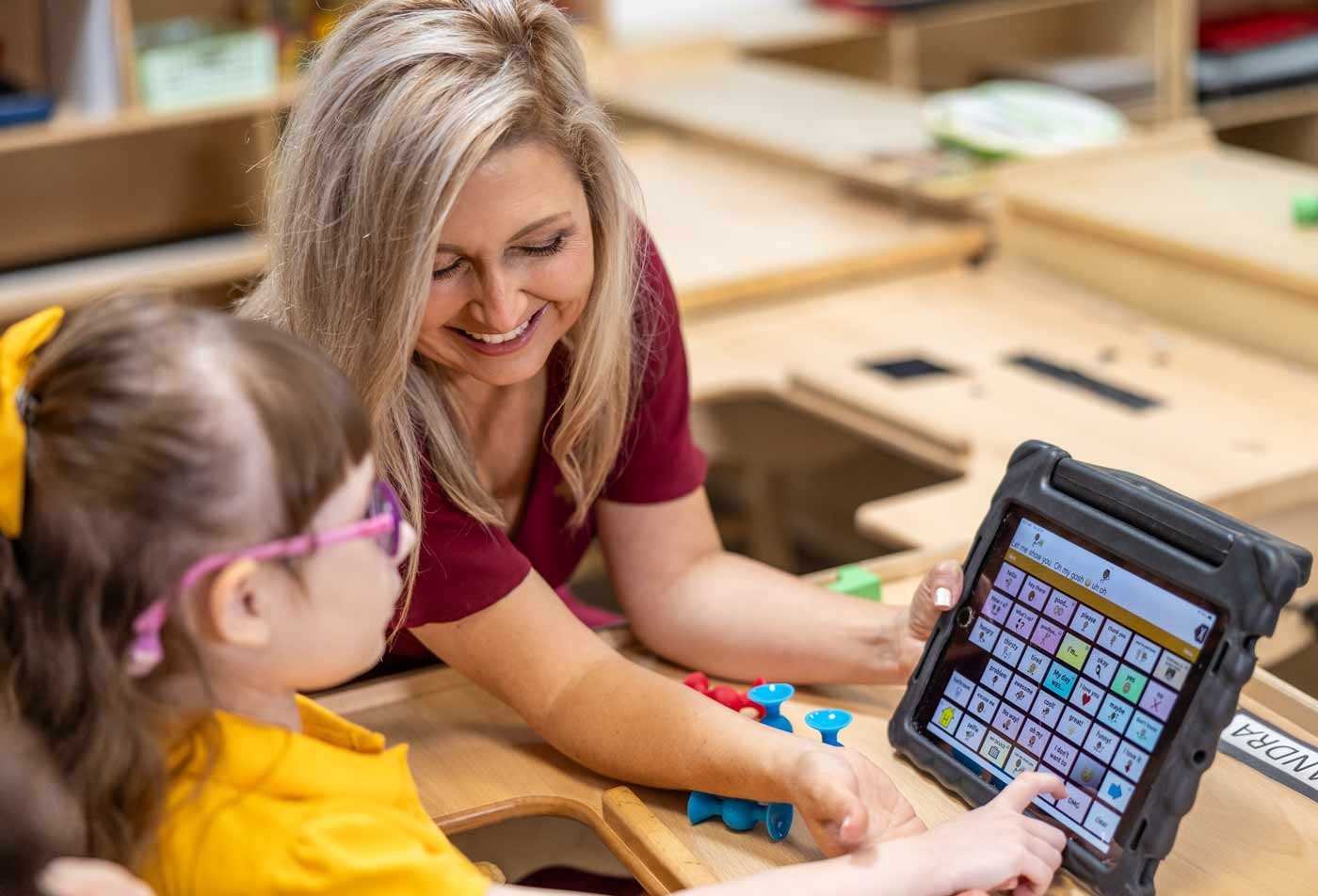 A teacher and a young girl interact in a classroom, focused on an communciation tablet, demonstrating the skills gained in the MEd in Early Childhood Special Education (Teacher Certification).