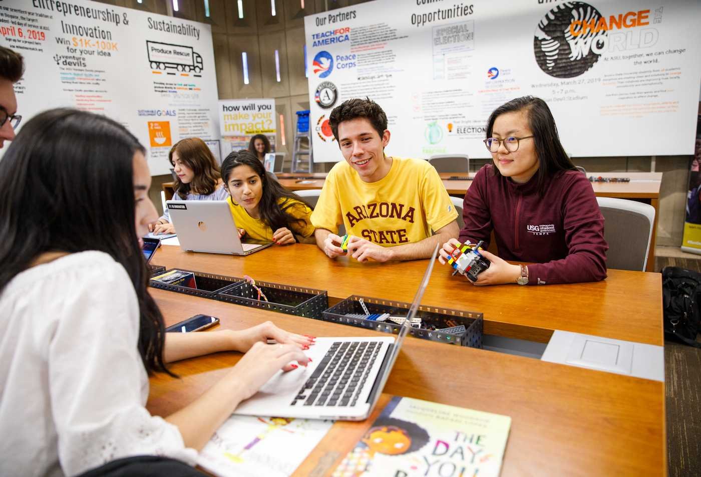 Students collaborate at a table in a workspace, working on laptops and holding devices, showcasing the collaborative learning environment of the minor in Educational Studies.