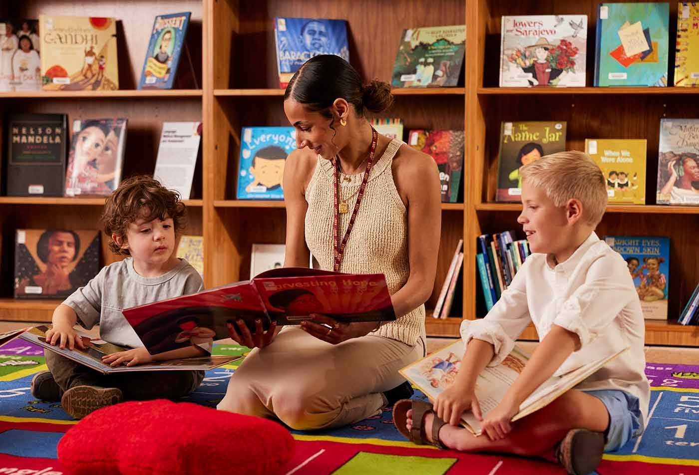 A woman reads a large picture book to two children sitting on a colorful rug in a library, demonstrating the expertise gained in the BAE in Elementary Education.