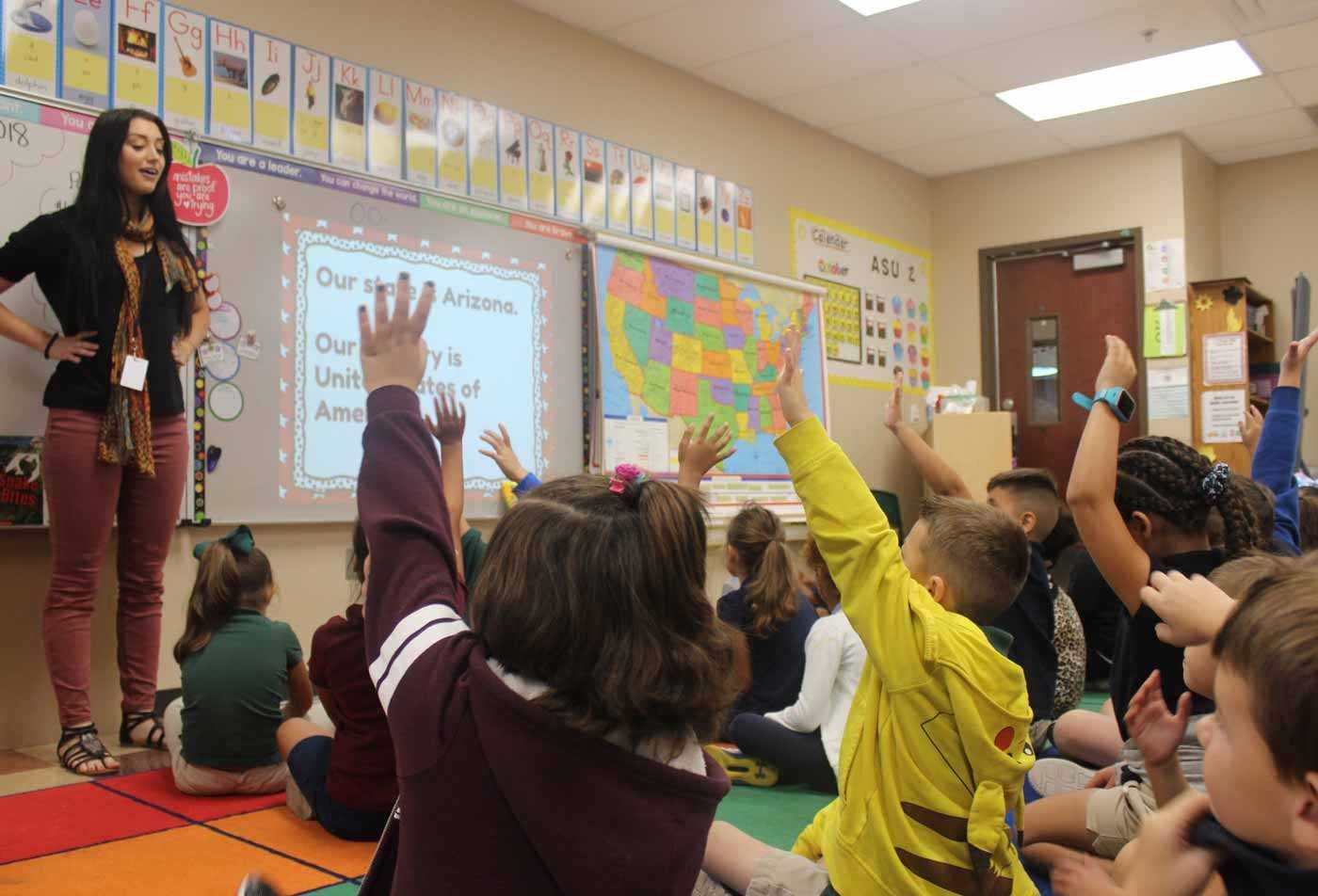 A teacher stands in front of a digital screen in a classroom, highlighting skills learned in the certificate in Elementary Education, with young students seated on a colorful carpet raising their hands to participate.