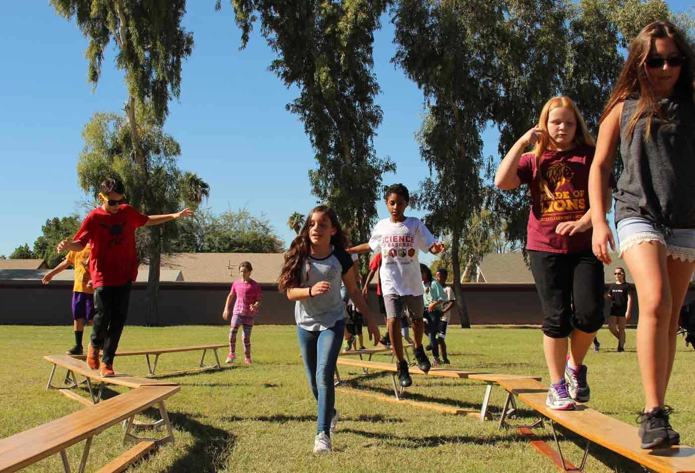 A group of children play on balance beams in a grassy area, showcasing the field explored in the BAE in Physical Education.